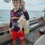 A boy stands on a boat holding a freshly caught fish with a gripper. The ocean and another person are visible in the background.