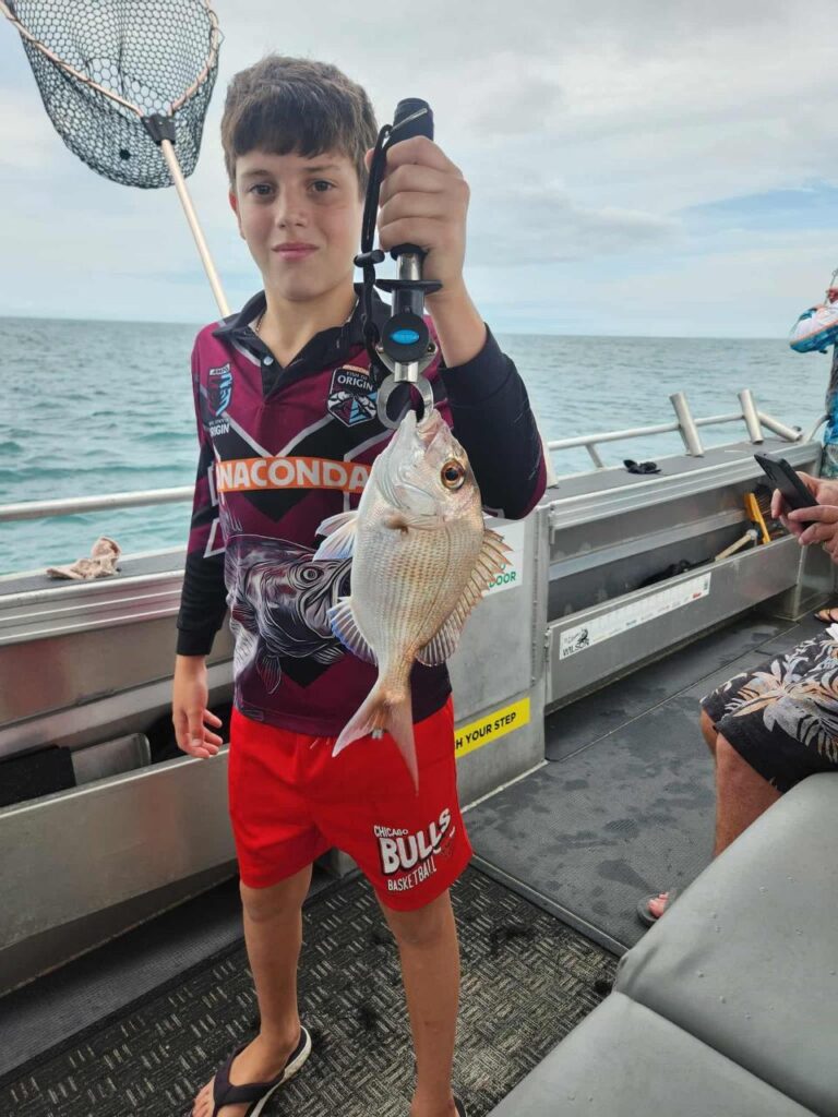 A boy stands on a boat holding a freshly caught fish with a gripper. The ocean and another person are visible in the background.