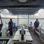 Four people, including two children, are fishing from a boat on calm water under a cloudy sky; safety and warning signs are visible inside the boat.