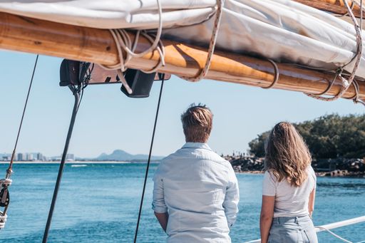 Two people stand on a boat, facing the water and distant shoreline, with sail rigging overhead and clear blue skies.