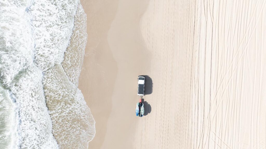 Aerial view of a white truck parked on a sandy beach near the shoreline, with surfboards or paddleboards attached to its rear. Tire tracks and waves are visible.