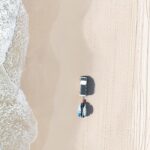 Aerial view of a white truck parked on a sandy beach near the shoreline, with surfboards or paddleboards attached to its rear. Tire tracks and waves are visible.