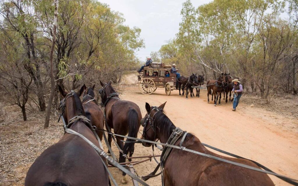 A group of horses pulling a mail stagecoach on a dirt road, with riders and handlers nearby among dry trees and brush.