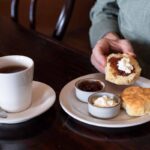 A person spreads cream and jam on a biscuit, with a cup of tea, extra jam, and cream on a plate on a wooden table.