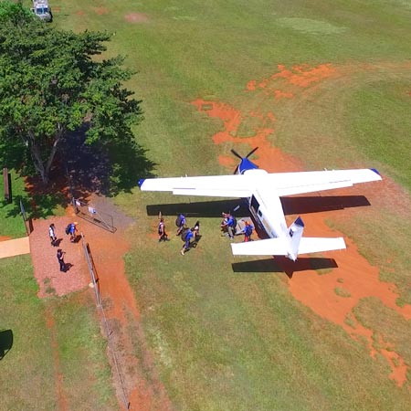 A small plane is parked on a grassy field as several people walk from the aircraft toward a nearby fence and trees.