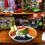 A plate of assorted fruits and vegetables sits on a counter surrounded by packaged food items, gift baskets, and condiments on shelves in a store.