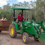 A man drives a green tractor pulling a wooden trailer filled with children through a rural, wooded area on a dirt path.