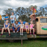 A group of children and one adult sit and stand on an old flatbed truck labeled “Water Ridge Farm” in an outdoor setting with trees and grass.