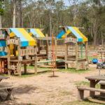 Outdoor playground with wooden play structures, colorful roofs, toy cars, picnic tables, and trees in the background.