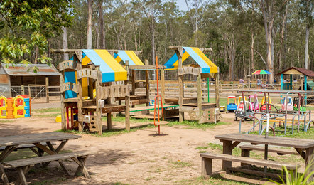 Outdoor playground with wooden play structures, colorful roofs, toy cars, picnic tables, and trees in the background.