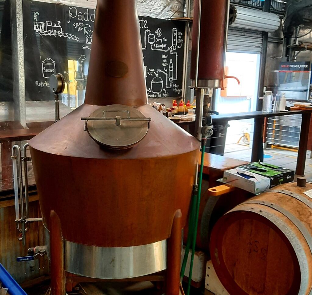 Large copper brewing kettle with a round hatch on top, situated in a brewery with a wooden barrel and chalkboard menu in the background.