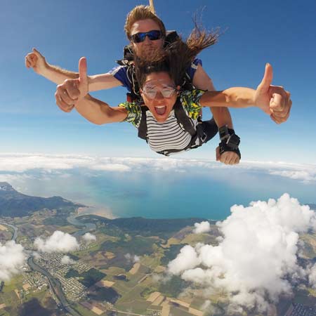 Two people tandem skydiving above a landscape with fields, river, and coastline visible below, both giving thumbs-up gestures.