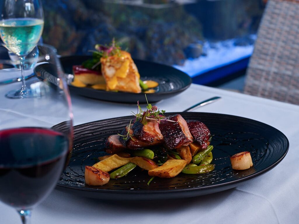 A plated gourmet meal with roasted meat, potatoes, and vegetables on a black dish, accompanied by glasses of red and white wine on a white tablecloth.