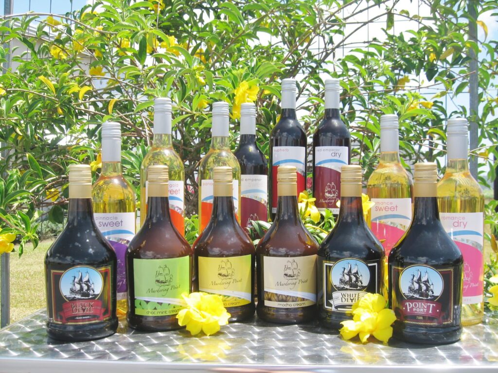 Assorted bottles of wine and liqueur are displayed on a metal table outdoors, with yellow flowers and green foliage in the background.