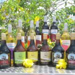 Assorted bottles of wine and liqueur are displayed on a metal table outdoors, with yellow flowers and green foliage in the background.