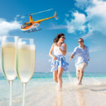 A man and woman walk along a beach shoreline, smiling, with a helicopter flying overhead and two glasses of champagne in the foreground.