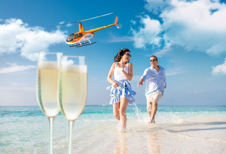 A man and woman walk along a beach shoreline, smiling, with a helicopter flying overhead and two glasses of champagne in the foreground.