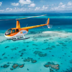 A yellow and orange helicopter flies over clear turquoise water and coral reefs near a small sandbar under a partly cloudy sky.