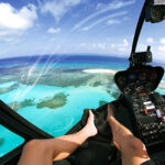 View from inside a helicopter cockpit flying over clear blue ocean water and coral reefs, with a person's bare legs visible on the seat.
