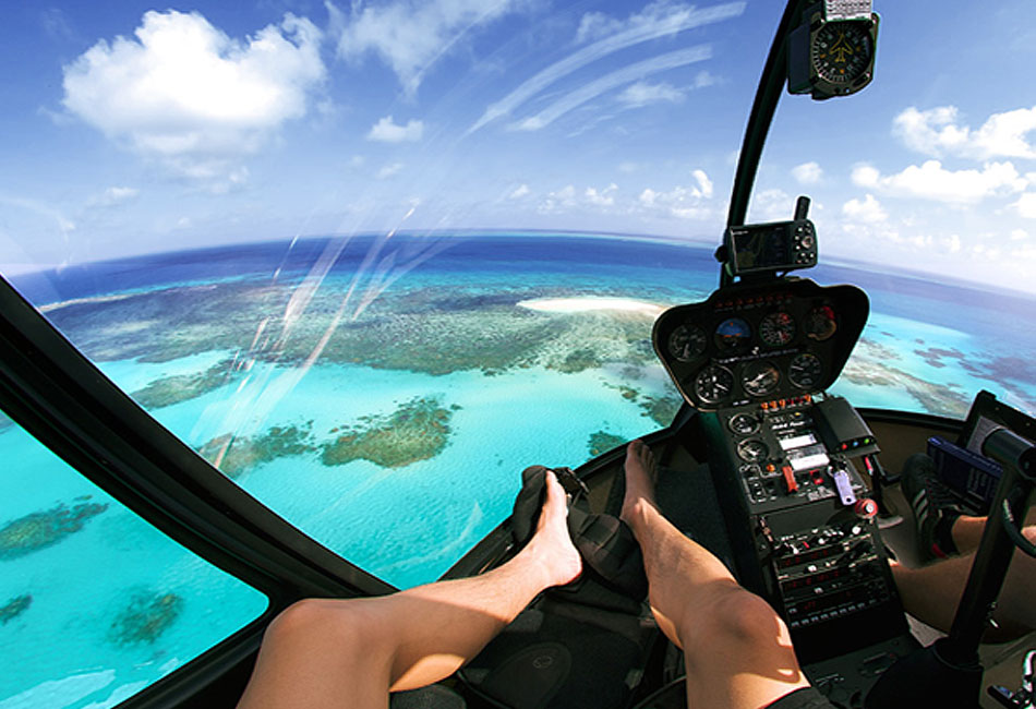 View from inside a helicopter cockpit flying over clear blue ocean water and coral reefs, with a person's bare legs visible on the seat.