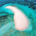 Aerial view of a small, crescent-shaped sandbar surrounded by clear turquoise water and shallow coral reefs.