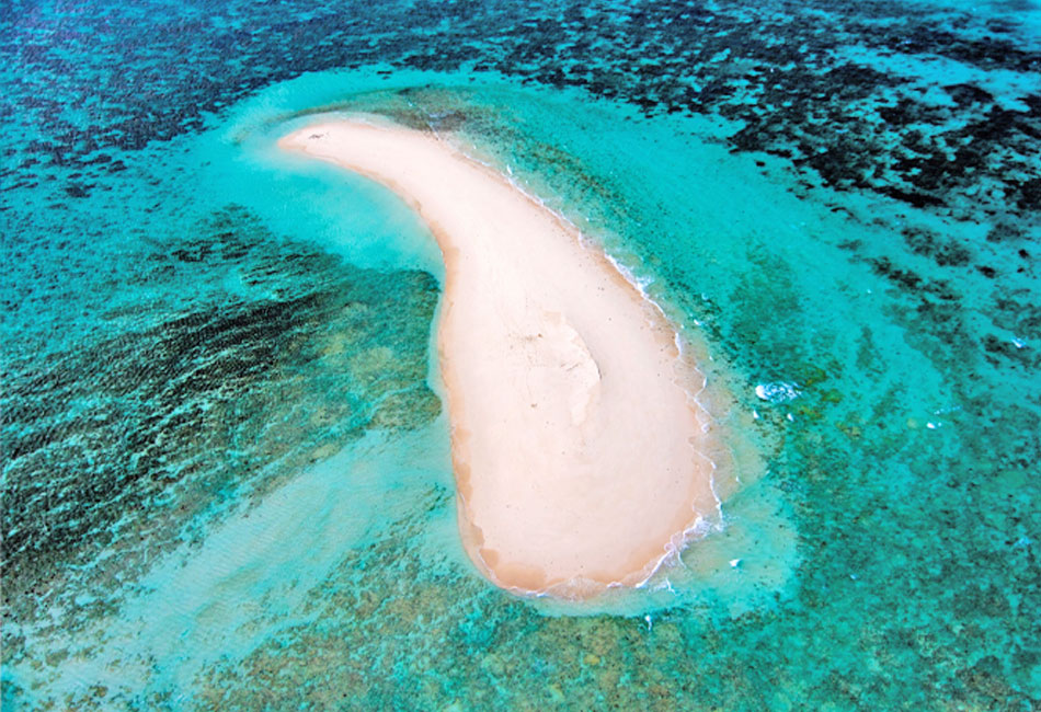 Aerial view of a small, crescent-shaped sandbar surrounded by clear turquoise water and shallow coral reefs.