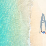 Aerial view of two people lying on a sandy beach near clear turquoise water, with their shadows forming the shape of a heart.