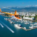 A bright orange helicopter flies over a marina filled with yachts and boats, with a city, harbor, and mountains visible in the background under a clear blue sky.