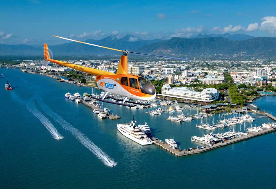 A bright orange helicopter flies over a marina filled with yachts and boats, with a city, harbor, and mountains visible in the background under a clear blue sky.