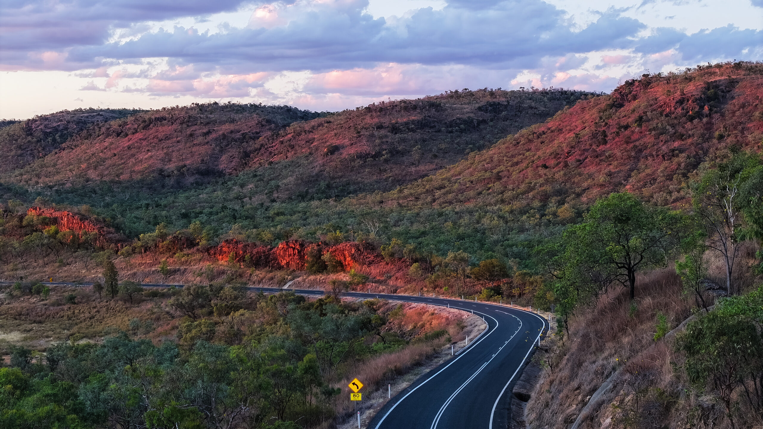 A winding paved road curves through a hilly landscape with green vegetation and red-tinged hills under a partly cloudy sky at sunset.