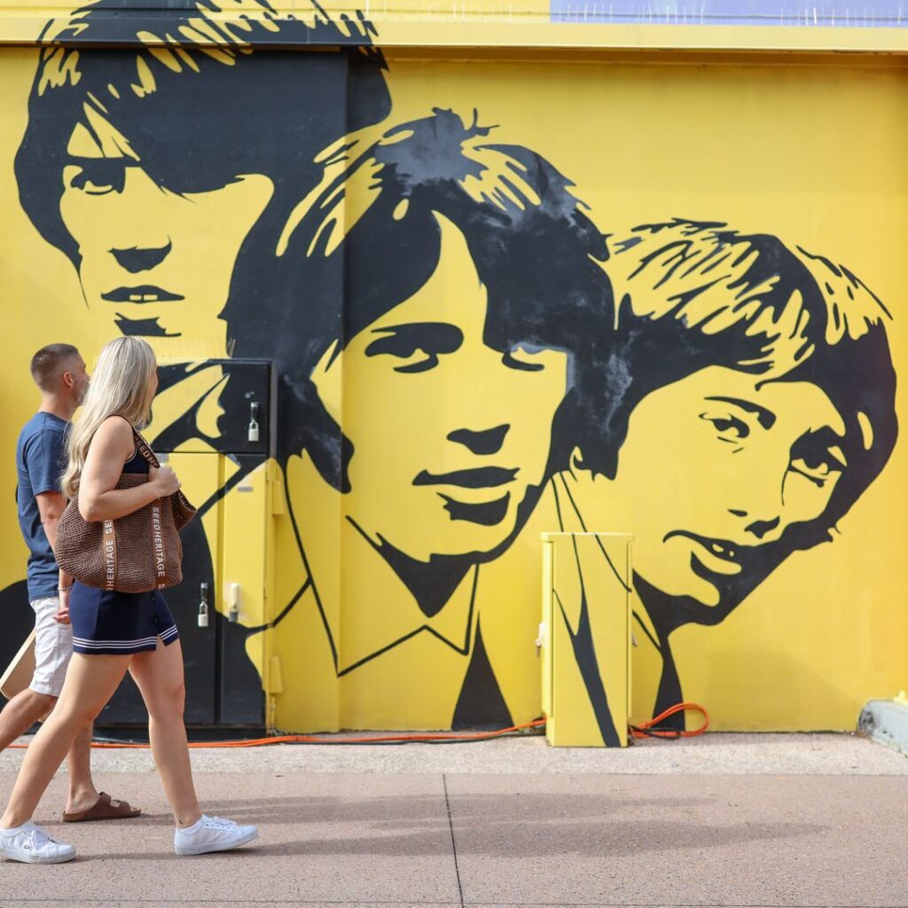 Two people walk past a yellow wall featuring a large black mural of three stylized faces in a graphic art style.