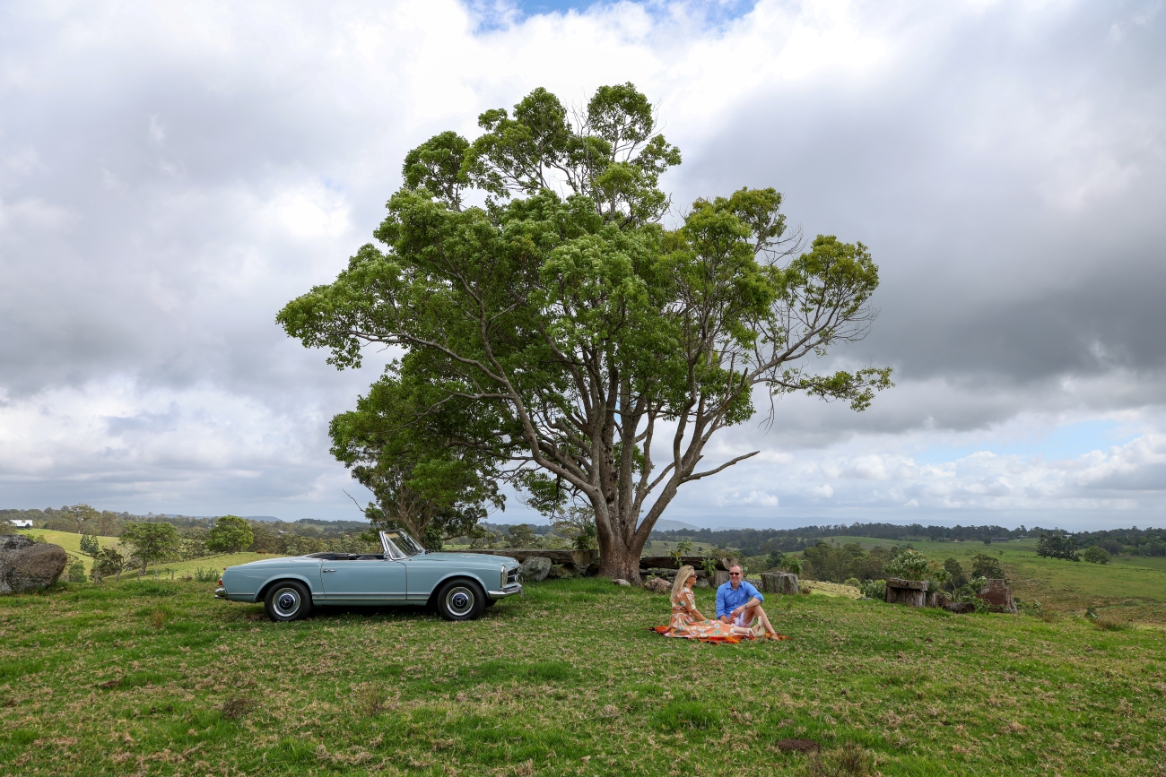 A blue convertible car is parked on grass near a large tree while two people sit on a blanket having a picnic under the tree, with a countryside landscape in the background.