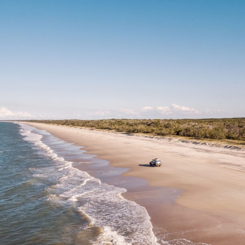 A vehicle drives along a wide, empty sandy beach next to the ocean, with a line of green vegetation in the background under a clear sky.