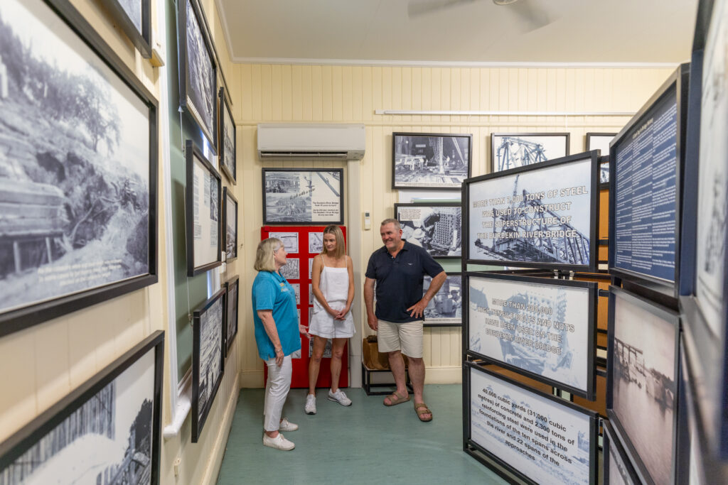 Three adults stand and talk inside a small museum room lined with framed historical photographs and informational displays.