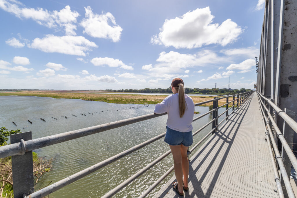 A person in a white shirt and denim shorts stands on a bridge, looking out over a river under a partly cloudy sky.