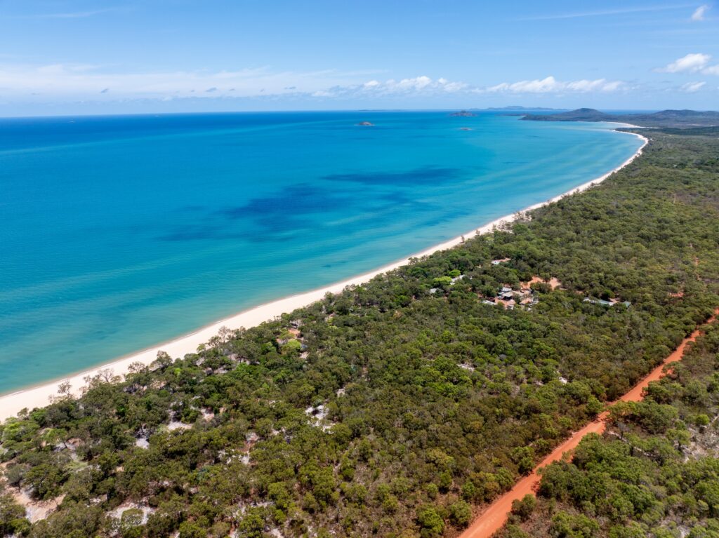 Aerial view of a long sandy beach bordering clear turquoise water, with dense green forest and a red dirt road running parallel to the shoreline.