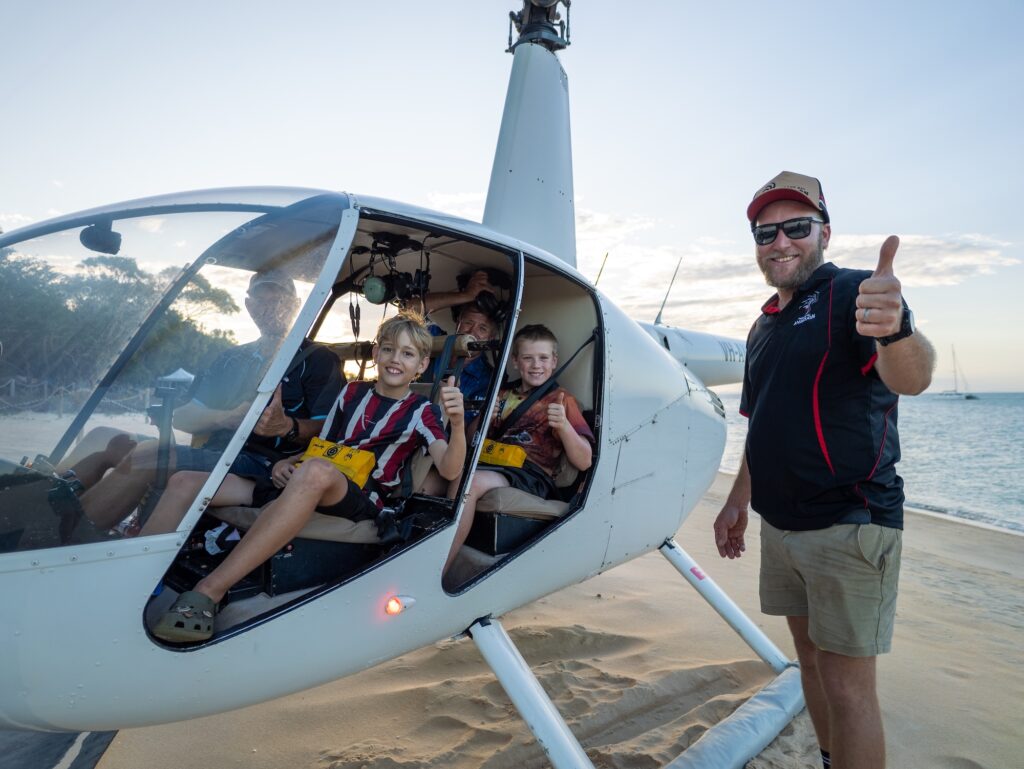 Three children sit inside a small helicopter on a sandy beach, smiling and giving thumbs up, while an adult stands outside giving a thumbs up.