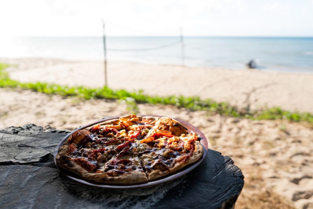 A pizza on a plate sits on a wooden surface with a sandy beach and ocean in the background.