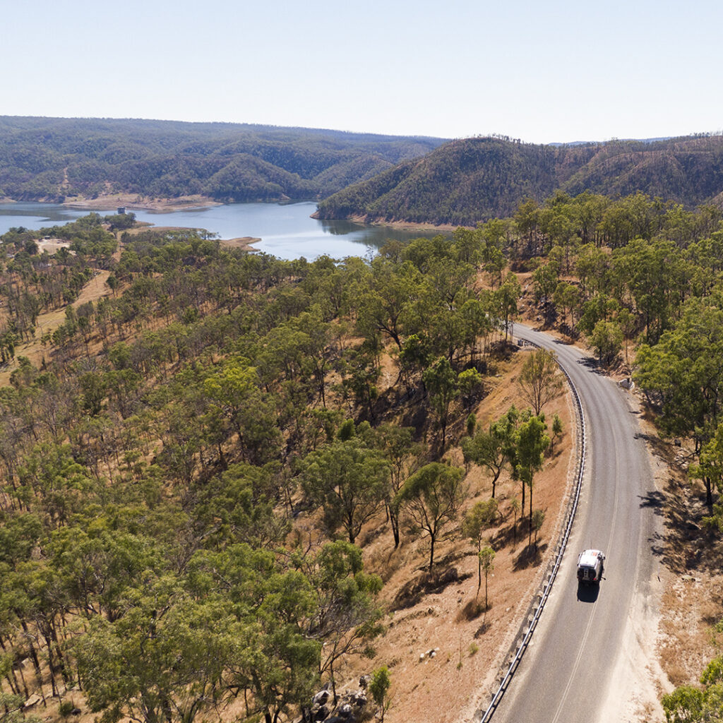 A car towing a caravan drives along a winding road beside a lake, surrounded by dry, tree-covered hills under a clear sky.