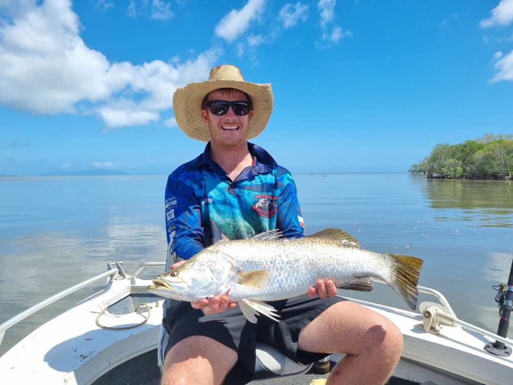 Person wearing a straw hat and sunglasses sitting on a boat, holding a large fish with both hands, with calm water and blue sky in the background.