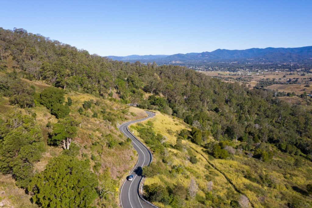 A winding road curves through green hills with a single car driving; trees cover one slope, and a valley with distant mountains is in the background under a clear blue sky.