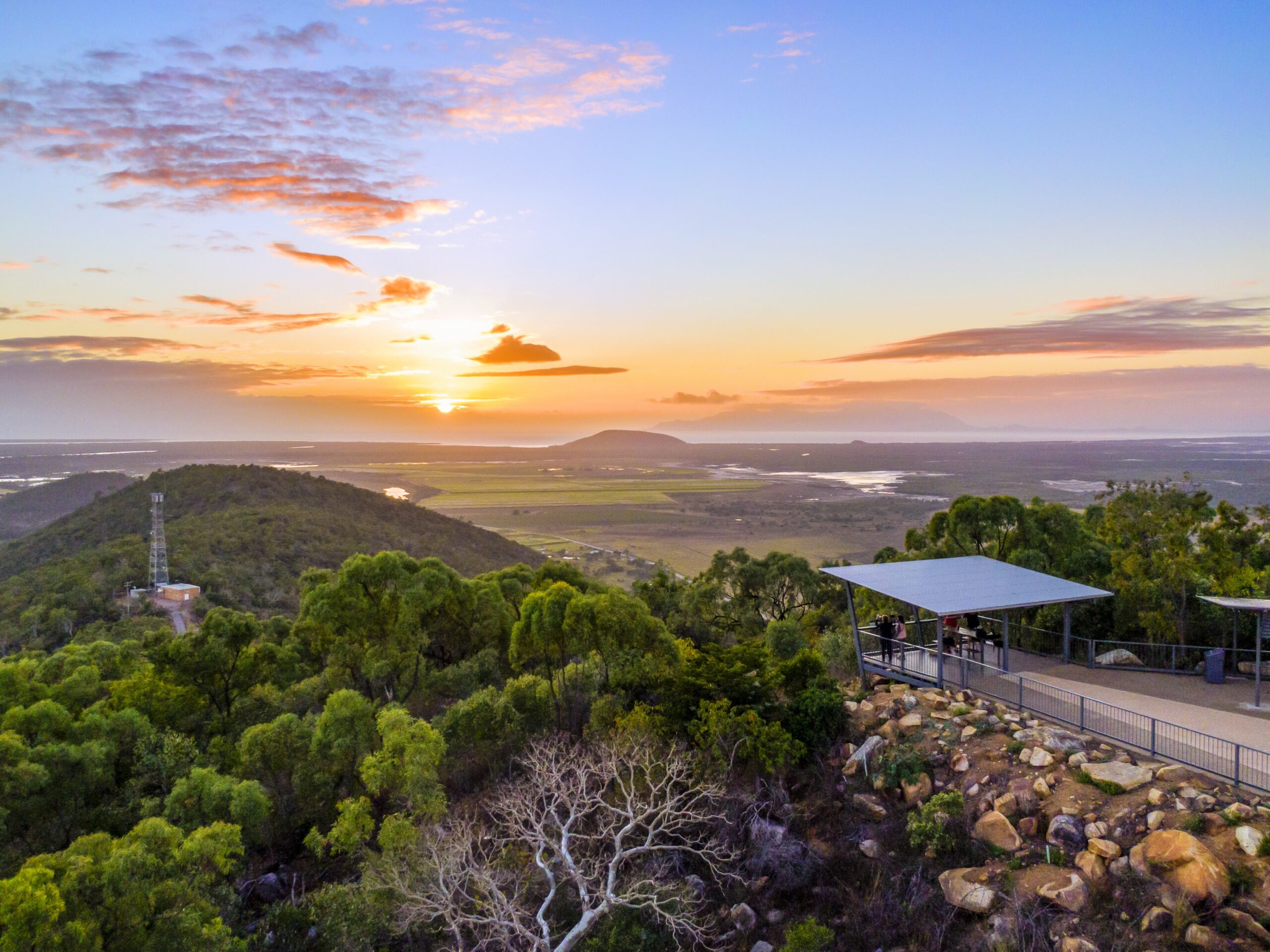 A scenic overlook with a shelter and lush greenery, facing a sunrise over hills, fields, and distant mountains under a partly cloudy sky.