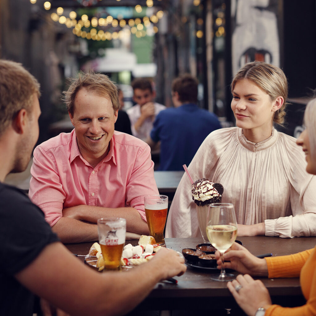 Four people sit at an outdoor table with drinks and desserts, talking and smiling under string lights in an urban setting.