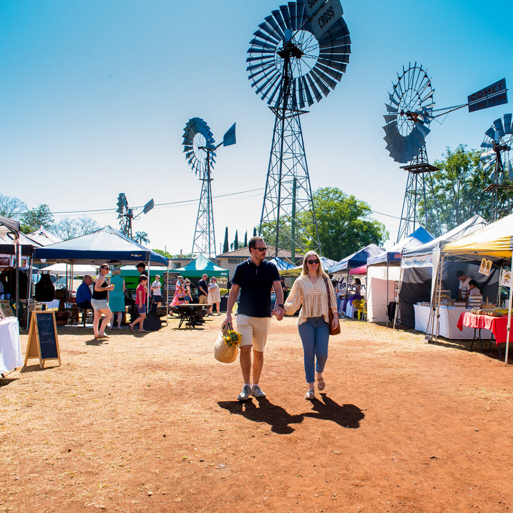 A couple holding hands walks through an outdoor market with windmills in the background and various vendor tents around them under a clear blue sky.