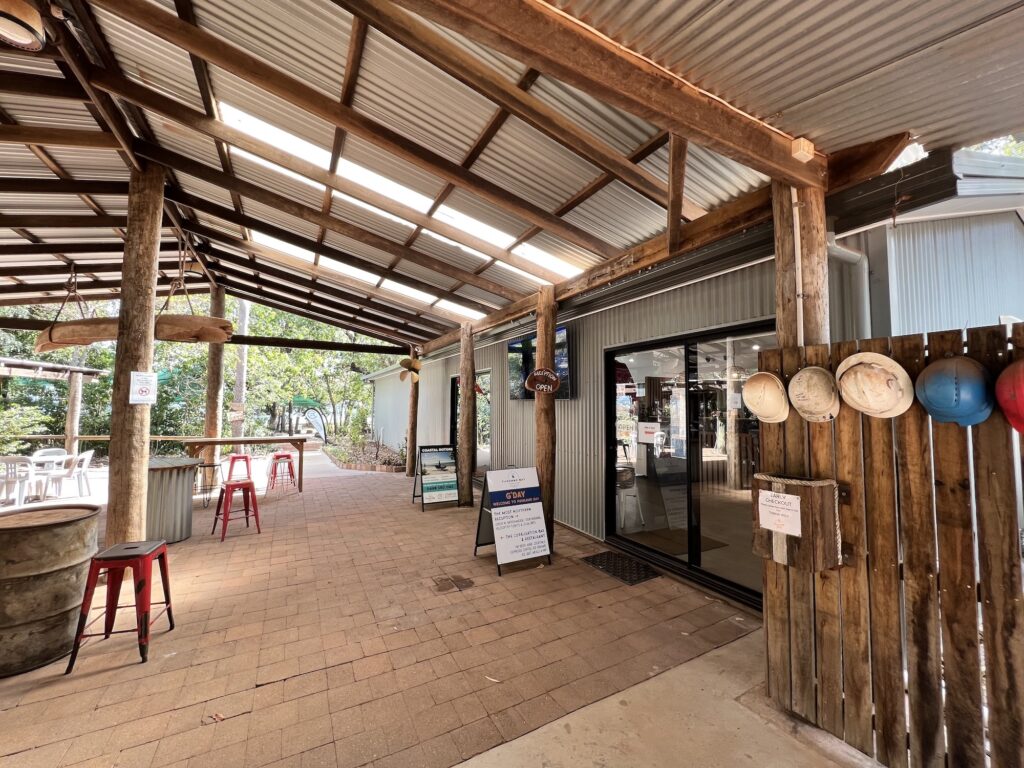 Outdoor seating area with a corrugated metal roof, wooden beams, red stools, a glass door, and various signs and hats hanging on a wooden fence.