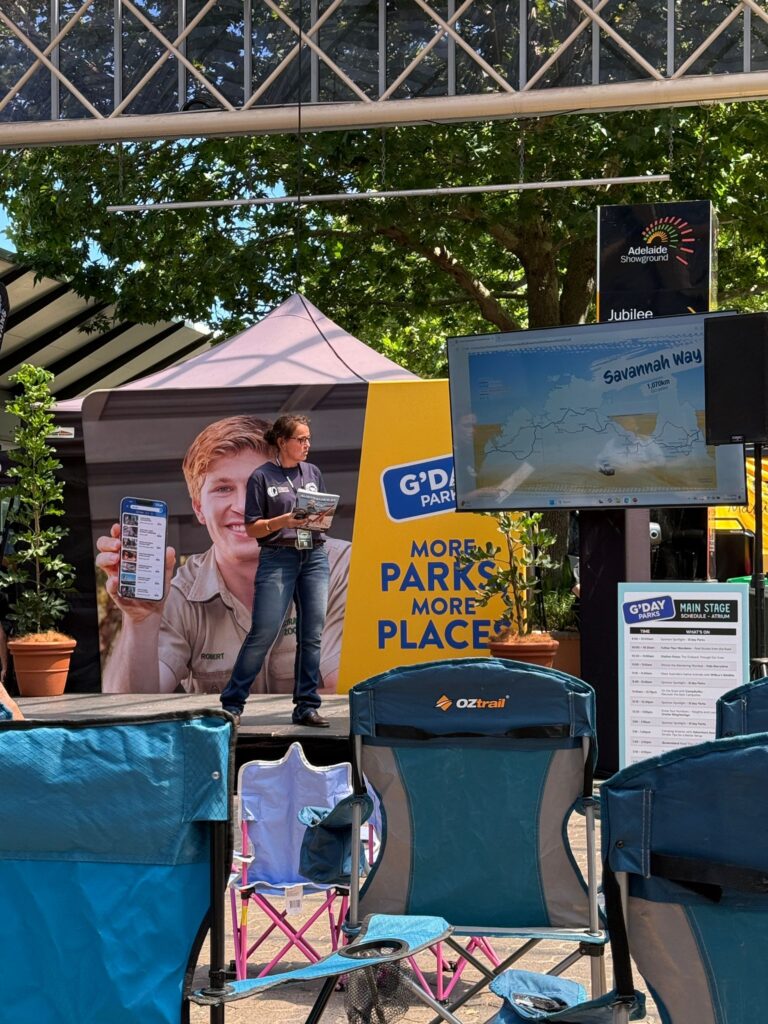 A person stands on an outdoor stage with a microphone and papers, beside a large display promoting parks. A TV screen shows a map, and empty chairs are in the foreground.