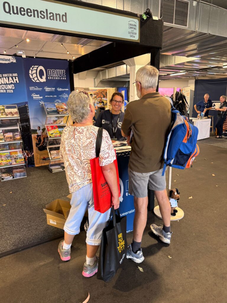 Two people talk to a staff member at the Queensland tourism booth, surrounded by brochures and promotional materials in an indoor exhibition setting.