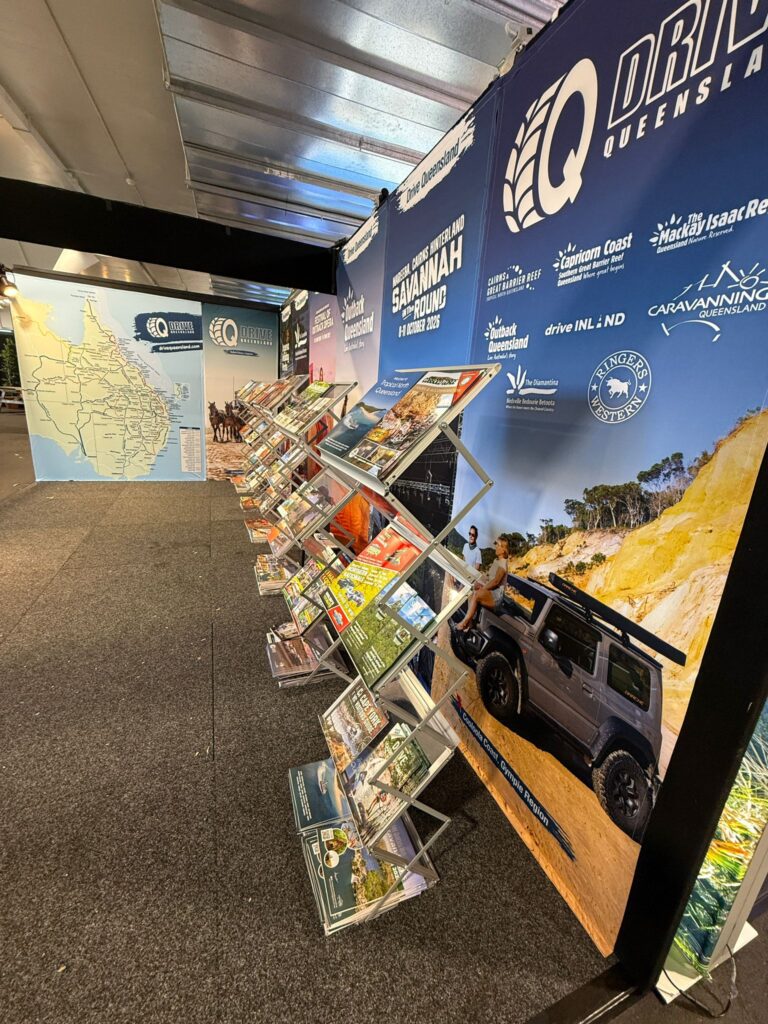A display of travel brochures and maps for Queensland, Australia, arranged on racks at an indoor tourism information booth.