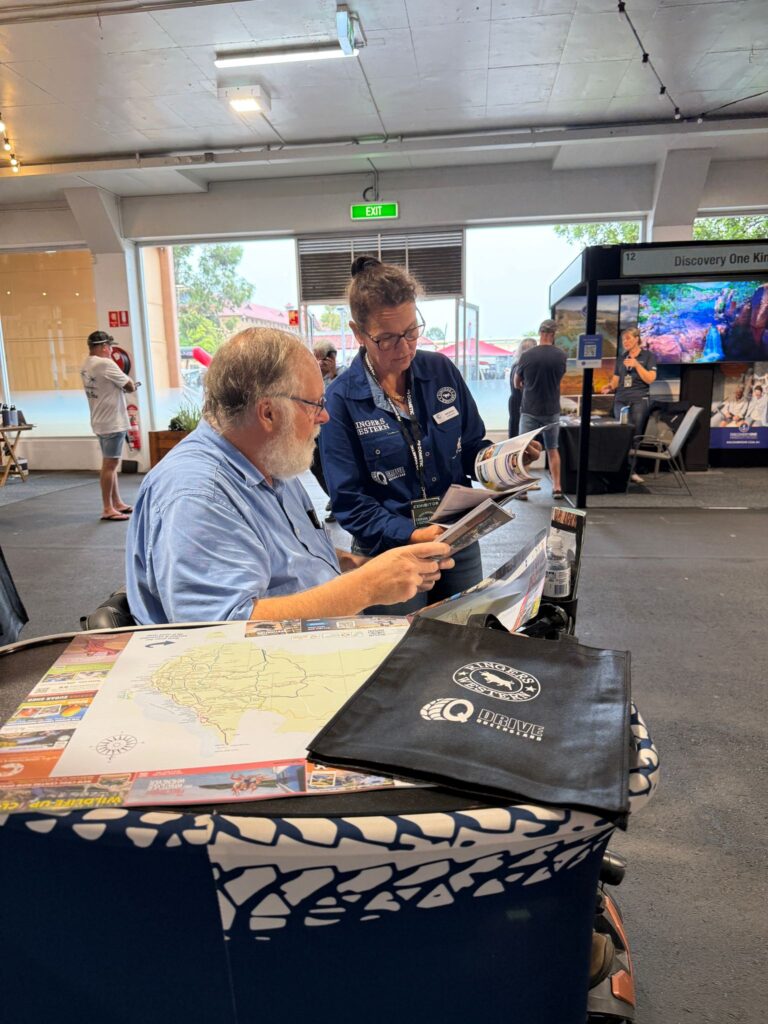 Two people at an indoor event look at brochures and maps on a table. A branded tote bag is in the foreground. Other attendees and displays are visible in the background.