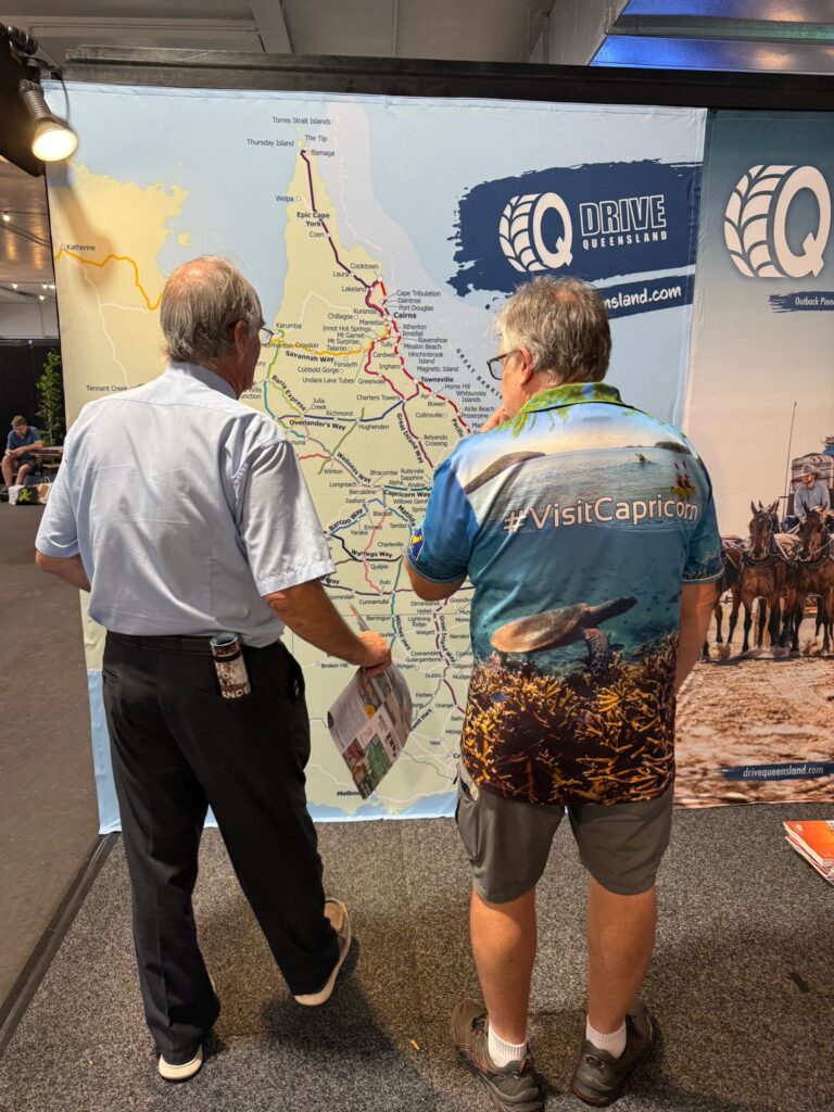 Two men stand in front of a large map of Queensland, Australia; one points at the map while the other wears a 